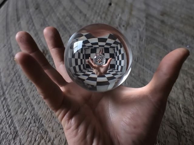 An open hand holding a clear glass sphere that reflects a chessboard pattern and an infinite optical effect of the same hand.